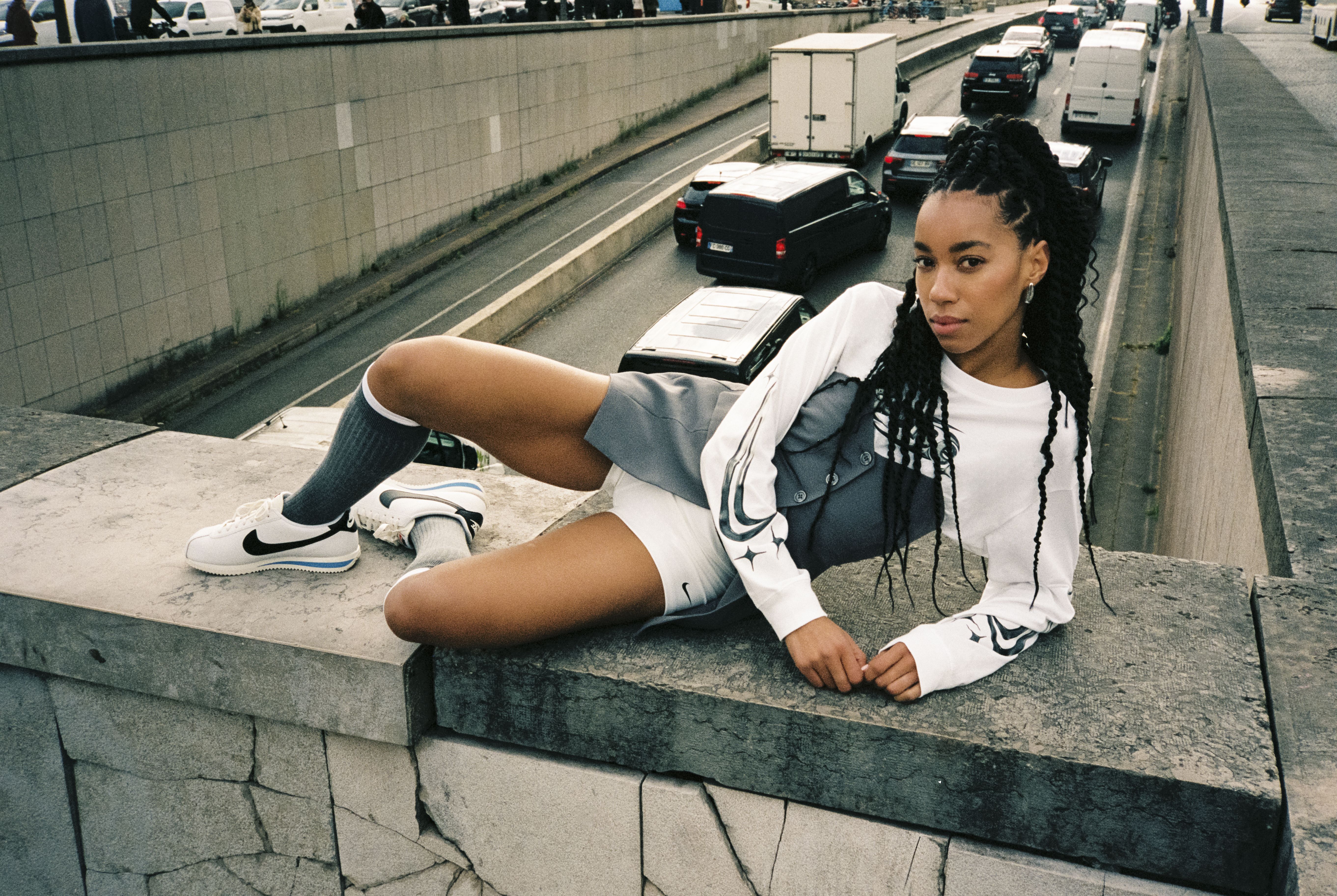 Ženska v beli NIKE majici in belih NIKE čevljih pozira na betonskem zidu nad prometno cesto.